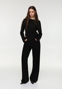 Fekete hosszú ujjú felső csomós részlettel a deréknál, bőszárú fekete nadrággal, enyhén fényes hatással és hegyes orrú cipőkkel párosítva.