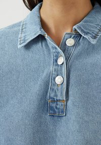 Camicia di jeans azzurro chiaro con colletto, tre bottoni a pressione bianchi e cuciture gialle a contrasto. Texture morbida, design informale.