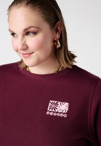 Bordeauxrode trui met een ronde hals, met een lichtroze grafisch ontwerp van bloemmotieven en golven op het linkerborstgebied.