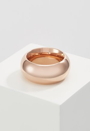 Gladde, brede roségouden metalen ring die rust op een wit voetstuk tegen een lichtgrijze achtergrond.