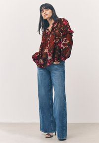 Blusa rossa con motivo floreale e maniche arricciate, abbinata a jeans blu a gamba larga e tacchi neri con punta aperta su sfondo neutro.