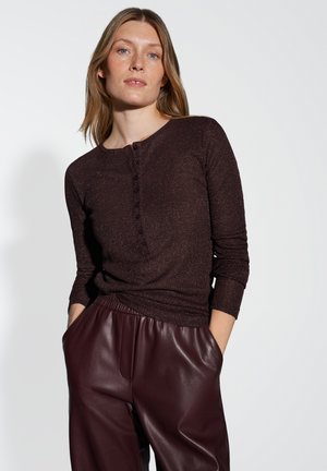 Vrouw met lichte huid en blond haar draagt een donkerbruin, knoopshirt met lange mouwen en bordeauxkleurige leren broek met haar handen in de zakken.