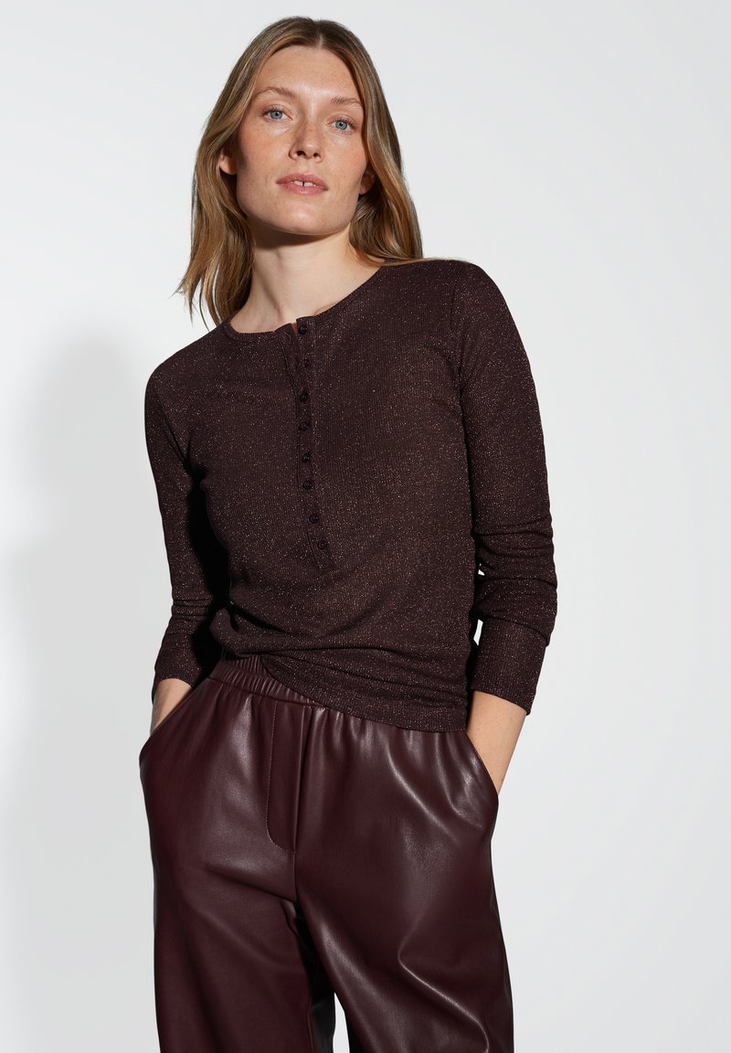 Una donna con pelle chiara e capelli biondi indossa una maglia lunga a maniche con bottoni di colore marrone scuro e pantaloni in pelle bordeaux, con le mani nelle tasche.