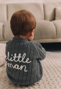 Pull en tricot gris à texture côtelée, avec le texte « petit homme » en lettres blanches au dos, porté par un enfant assis.