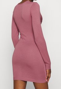 Žebrované šaty s dlouhým rukávem v světle růžové barvě, s přiléhavým střihem. Lem a manžety jsou jemně žebrované pro texturu.