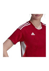 Rotes Fußballtrikot mit kurzen Ärmeln, einem strukturierten geometrischen Muster, weißen Akzenten und drei horizontalen weißen Streifen auf den Schultern.