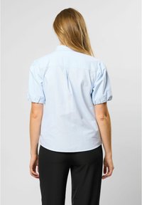 Light blue short-sleeve blouse with puffed sleeves, a structured back yoke, and a rounded hem, paired with black pants. Smooth cotton texture.