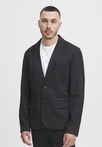 Black blazer made of soft fabric, single-breasted with two buttons, notched lapels, and two front pockets. Worn over a white t-shirt.