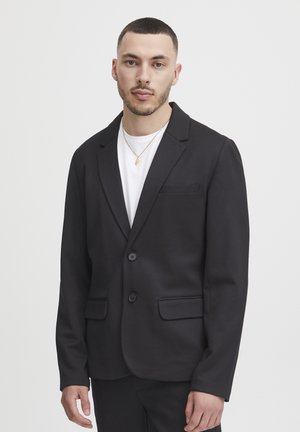 Black blazer made of soft fabric, single-breasted with two buttons, notched lapels, and two front pockets. Worn over a white t-shirt.