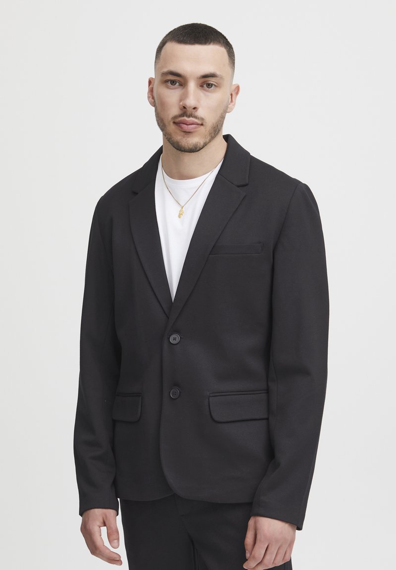 Black blazer made of soft fabric, single-breasted with two buttons, notched lapels, and two front pockets. Worn over a white t-shirt.
