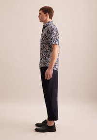 Camicia a maniche corte floreale nei colori blu navy, beige e bianco; abbinata a pantaloni scuri e sartoriali e scarpe nere, vista di lato.