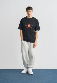 Μαύρο μπλουζάκι με κόκκινο και λευκό λογότυπο «Air Jordan», σε συνδυασμό με ανοιχτό γκρι φόρμα και λευκά αθλητικά παπούτσια. Το μοντέλο στέκεται σε μπλε χαλί.