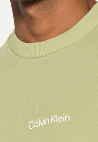 Světle zelené bavlněné tričko s krátkým rukávem a ribbovaným límcem, zdobené bílým logem "Calvin Klein". Hladká textura a přiléhavý design.