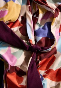 Bluse aus Blumenstoff mit Knotendetail in einem kräftigen lila Farbton, und lebhaften Farben wie Rot, Pink, Gelb und Blau in einer weichen, glatten Textur.