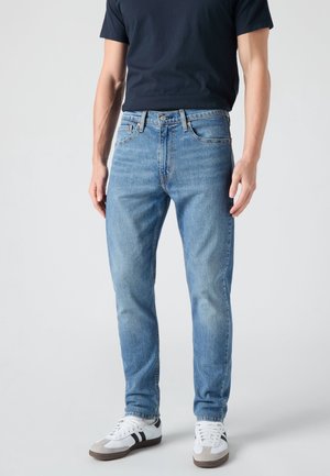 Homme portant un jean droit bleu clair, un t-shirt noir et des baskets blanches avec des rayures noires, debout sur un fond uni.