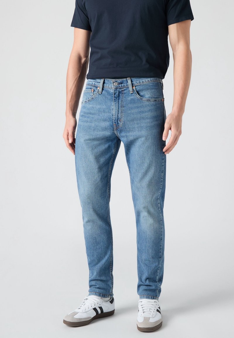 Homme portant un jean droit bleu clair, un t-shirt noir et des baskets blanches avec des rayures noires, debout sur un fond uni.