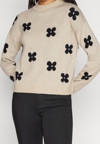 Beiger Strickpullover mit schwarzen Blumendesigns, hochgeschlossenem Kragen und lockerer Passform. Das Material wirkt weich und hat eine strukturierte Oberfläche.