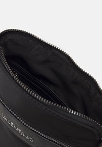 Sac en nylon noir avec une fermeture éclair, doté d'un intérieur texturé et d'un accent de logo. Le bord présente une finition de zip lisse pour plus de durabilité.