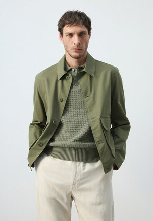Hombre con chaqueta abotonada verde oliva sobre suéter de punto texturizado y pantalones beige claros, de pie con las manos en los bolsillos contra un fondo liso.