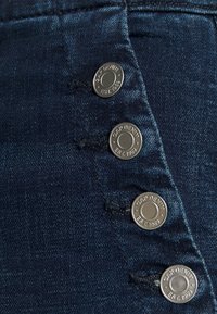 Mörk denimtyg med en slät textur som har fyra silverfärgade metallknappar präglade med "GAP DENIM EST. 1969" för stängning.