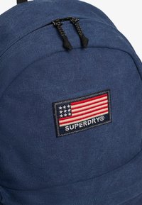 Niebieski plecak z materiału z zamknięciem na górze na zamek błyskawiczny, dwoma czarnymi uchwytami oraz naszywką z motywem amerykańskiej flagi i napisem "SUPERDRY".