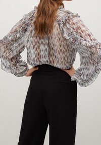 Donna con lunghi capelli castani che indossa una blusa semi-trasparente a fantasia con volant, infilata in pantaloni neri a vita alta, mani posate sui fianchi.