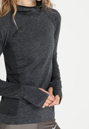 Kvinde iført en langærmet mørkegrå sportstop med tommelfingerhuller og teksturerede sidepaneler, hænderne foldet ved taljen.