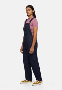 Dark denim overalls with adjustable shoulder straps, pockets, and yellow stitching, paired with a pink fitted t-shirt and yellow sneakers.