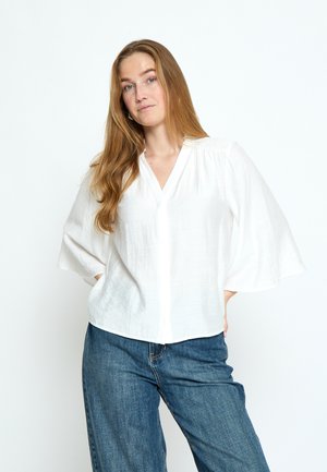 Junge Frau mit langen roten Haaren, die eine lockere weiße Bluse und blaue Jeans trägt, steht mit den Händen in den Taschen vor einem weißen Hintergrund.