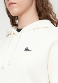 Sweat à capuche crème avec un petit logo de chaussure Converse en noir et blanc. Tissu texturé avec cordons de serrage et coupe décontractée.