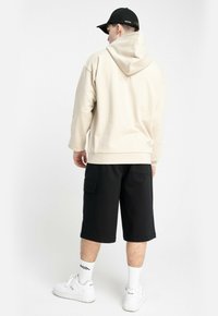 Mężczyzna stoi tyłem, ubrany w beżowy hoodie, czarne spodenki cargo, białe skarpetki, białe sneakersy i czapkę w kolorze czarnym.