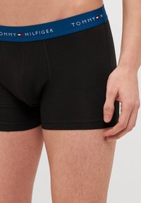 Musta puuvillainen boxer- alusvaate, jossa on sininen joustava vyötärö, jossa on valkoinen "TOMMY HILFIGER" -brändäys ja vaakasaumatiedot.