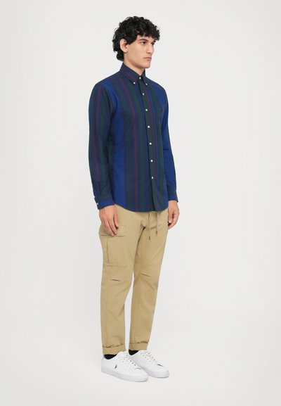Camisa abotonada de color azul oscuro con rayas verticales verdes y moradas, combinada con pantalones cargo beige y zapatillas blancas. Cuello plano y mangas largas.