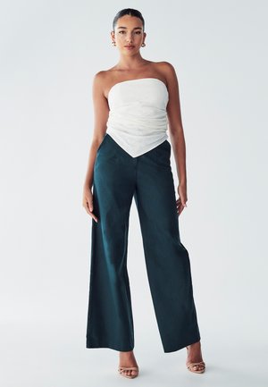 White strapless top with gathered texture; teal wide-leg pants; smooth fabric; neutral background; model stands confidently.