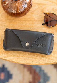 Zwarte leren brillenkoker gesloten met een zilveren knoop op een houten tafel naast een schildpaddenbril en een decoratief glazen voorwerp.