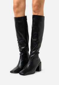 Bottes noires montant jusqu'au genou en cuir lisse, avec un bout carré, une finition texturée et un talon bloc épais. Dotées d'une fermeture éclair latérale.