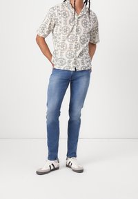 Kortärmad skjorta med knappslå i krämfärgad bas och svarta geometriska mönster, matchad med ljusblå slim fit-jeans och grå sneakers.