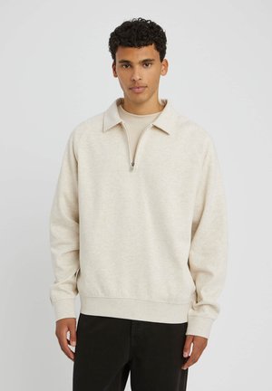 Sweatshirt beige à zip quart, avec un large col, des poignets côtelés et une texture douce, porté par-dessus un haut ajusté assorti et un pantalon sombre.