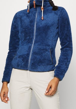 Vrouw met een blauwe, pluizige jas met rits en trekkoorden en lichtbeige broek, staand tegen een effen achtergrond.