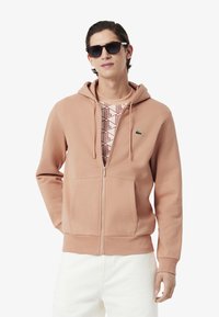 Sudadera rosa con cremallera hecha de algodón, presenta un bolsillo tipo canguro, puños acanalados y un pequeño logo verde en el pecho, llevada sobre una camisa con estampado.