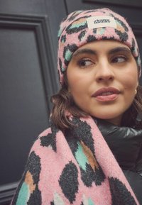Adīta rozā beanie cepure ar melnu, tirkīza un brūnu leopardu rakstu; logo uz priekšpuses. Mīksta, teksturēta šalle ar līdzīgu rakstu.