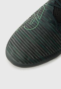 Svart atletisk sko med en texturerad yta med vertikala ränder och en grön accentdesign; lätt material för prestanda.