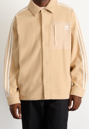 Man draagt een beige fleecejas met drukknopen, witte drievoudige strepen op de mouwen en een beige borstzak met Adidas-logo.