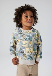 Lys gul genser med tegneseriestil dinosaurgrafikk i blått, grønt og grått, med ribbestrikkede mansjetter og kant. Kombinert med beige bukser.