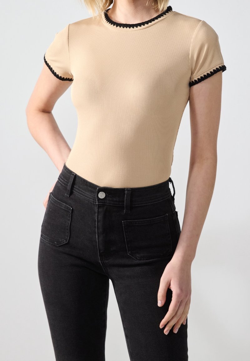 Femme portant un top beige côtelé ajusté à manches courtes avec bordure noire, associé à un jean noir taille haute avec des poches avant.