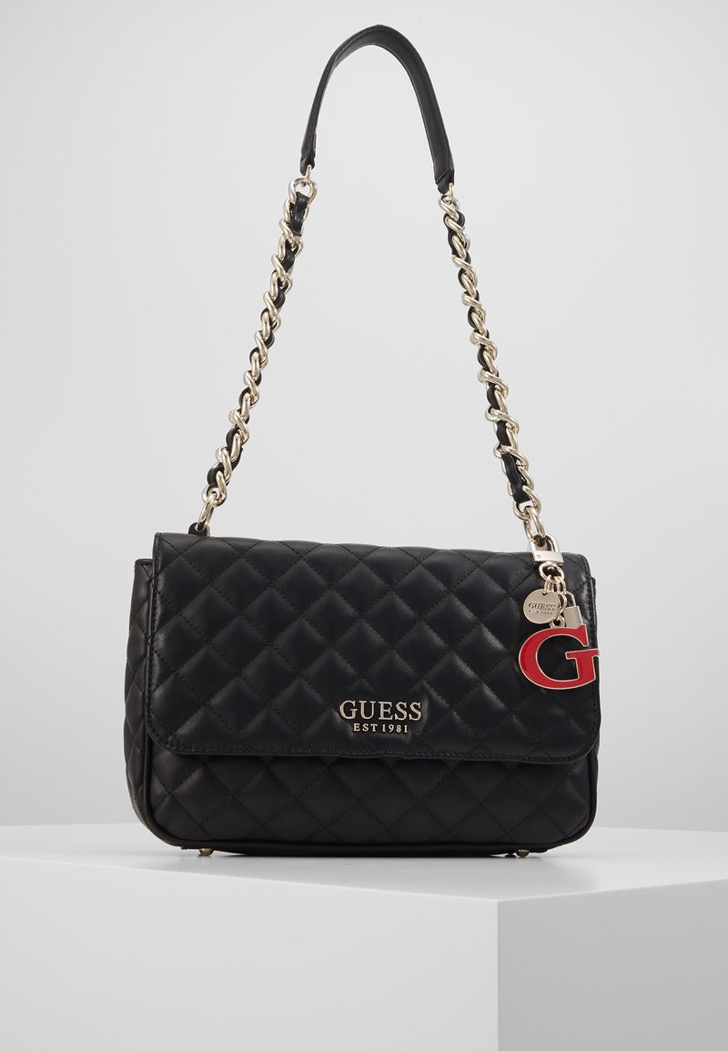 Sac à épaule matelassé noir avec bandoulière chaîne dorée, logo "GUESS EST 1981" et breloque rouge "G" sur le côté.