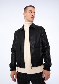 Veste en cuir noir avec une fermeture éclair à l'avant, deux poches sur la poitrine et des poignets côtelés, portée sur un col roulé en tricot crème.