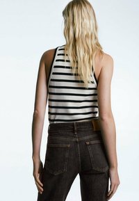 Top senza maniche a righe bianche e nere, abbinato a jeans a vita alta marrone scuro con tasche posteriori e etichetta in pelle sulla cintura.
