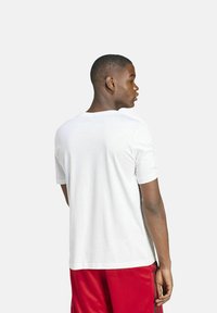 Camiseta blanca de manga corta con cuello redondo; textura suave y corte regular; llevada con pantalones cortos deportivos rojos, mostrando una vista trasera sencilla.