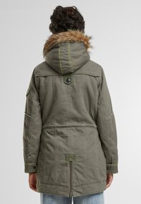 Olijfgroene parka met een met bont gevoerde capuchon, drukknoopdetails, zijzakken en zichtbare stiksels. Geweven stof met een gelaagd ontwerp.